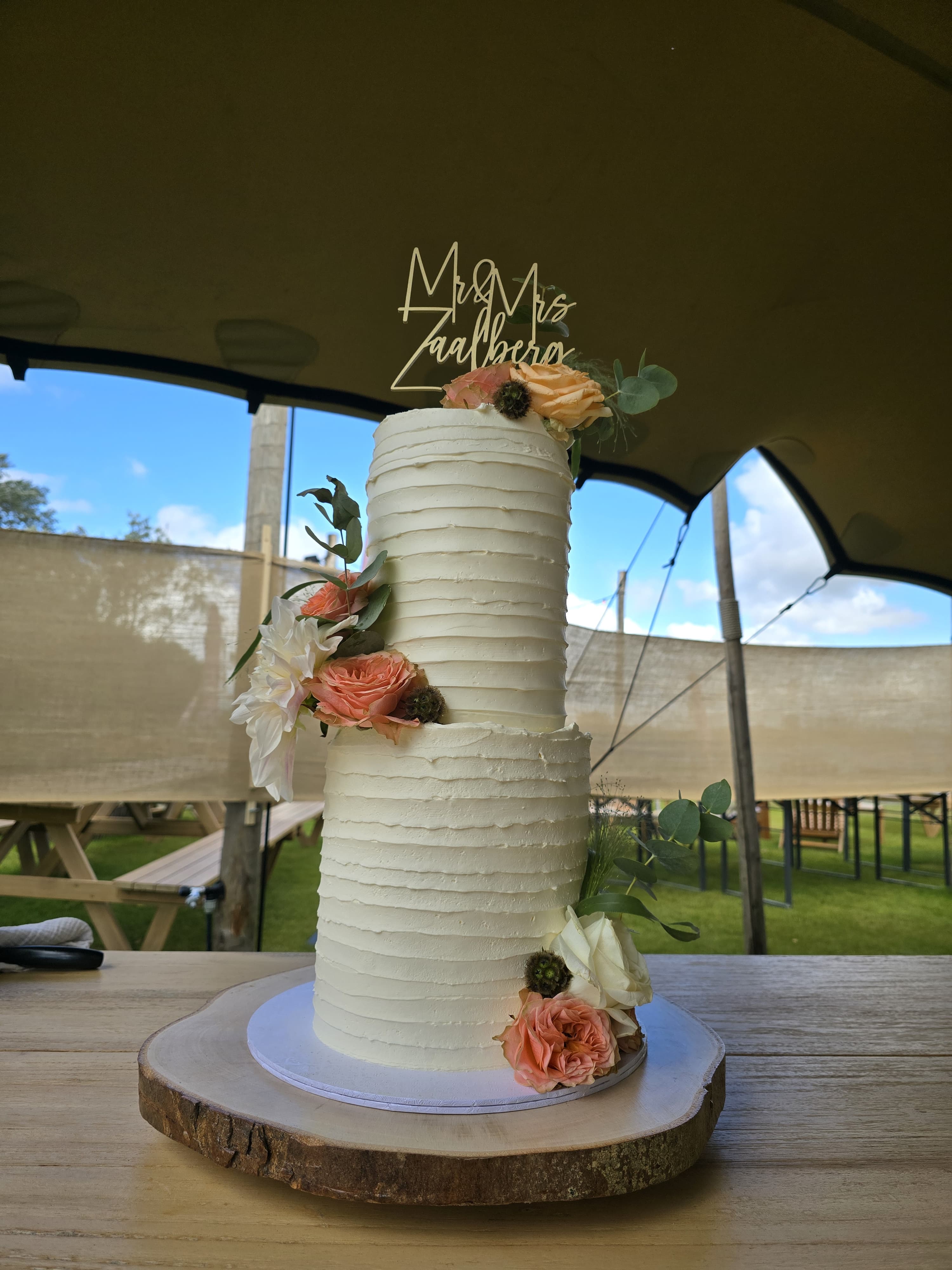 Klassieke hoge bruidstaart in botercrème met perzikkleurige rozen en eucalyptus op een houten plak